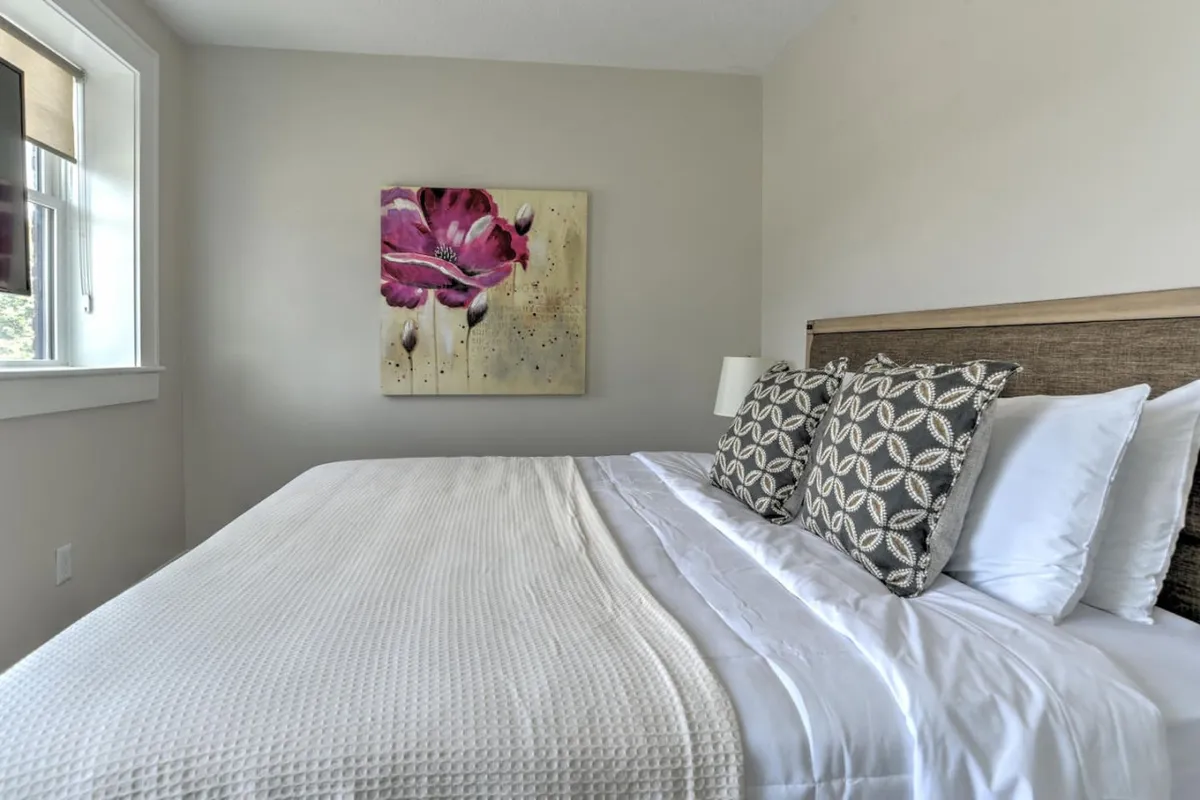 A tranquil bedroom featuring a bed with crisp white linens, tasteful artwork on the wall, and decorative pillows. Soft natural light filters in, creating a serene and inviting environment ideal for relaxation and restoration.