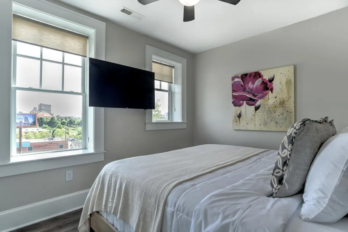 A quiet, well-appointed bedroom with a comfortable bed, stylish wall art, a television, and large windows for ample daylight. The neutral tones and uncluttered space evoke a sense of peace and privacy—ideal for a second bedroom retreat.