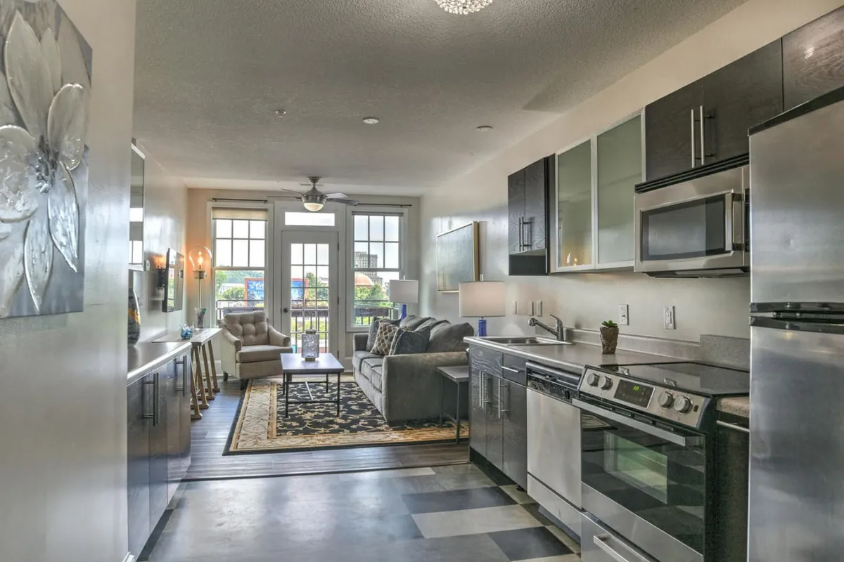 This image displays a cozy and contemporary open-plan living area, with a sofa, kitchen island, and dining area arranged in a light-filled space. The scene includes balcony doors that draw in natural light, highlighting the seamless flow between lounging, dining, and kitchen spaces.