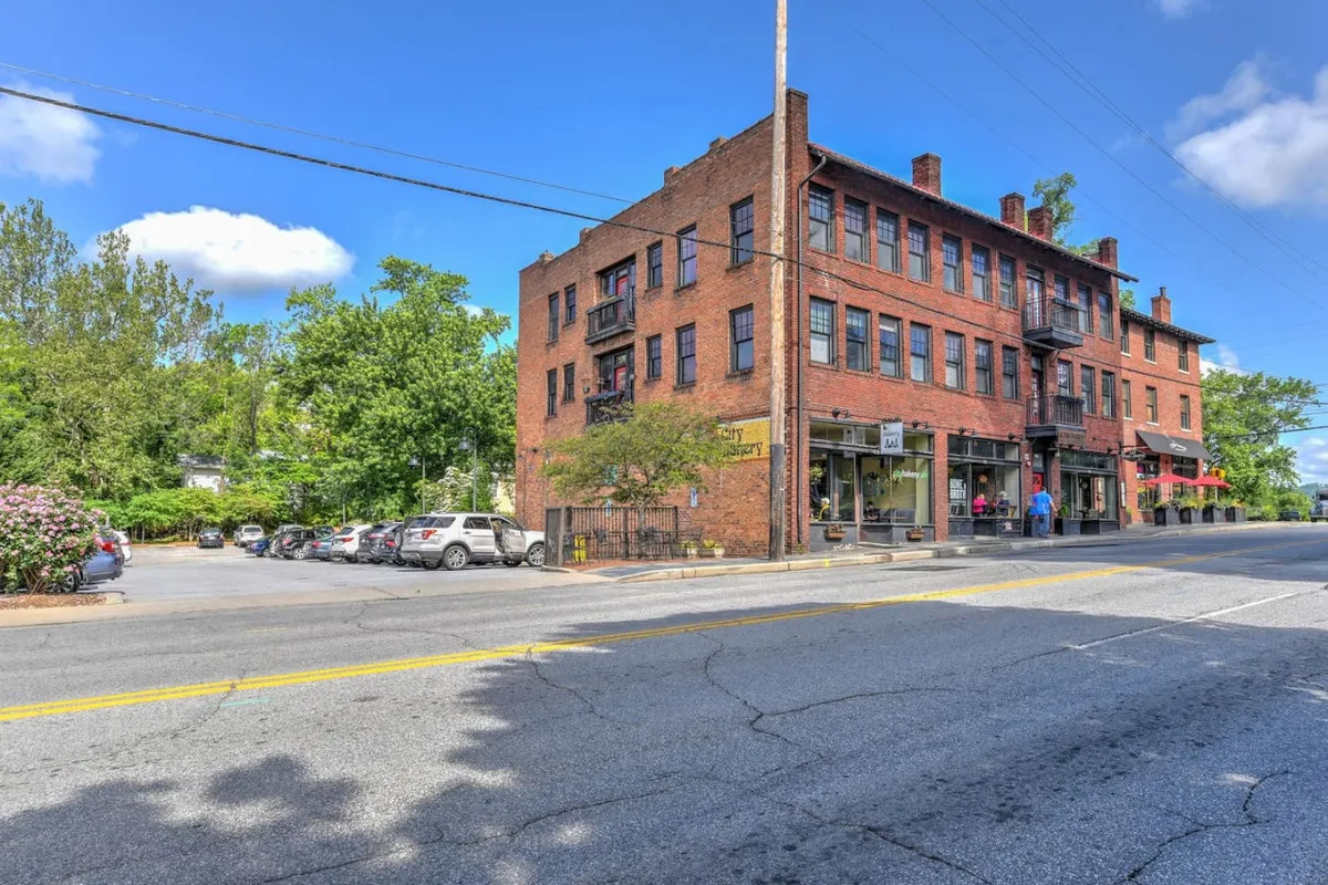 A bustling streetscape featuring a historic brick building with storefronts, green trees, and parked cars. The scene is lively with people strolling and outdoor café seating, showcasing the vibrant, community-oriented neighborhood just outside the condo.