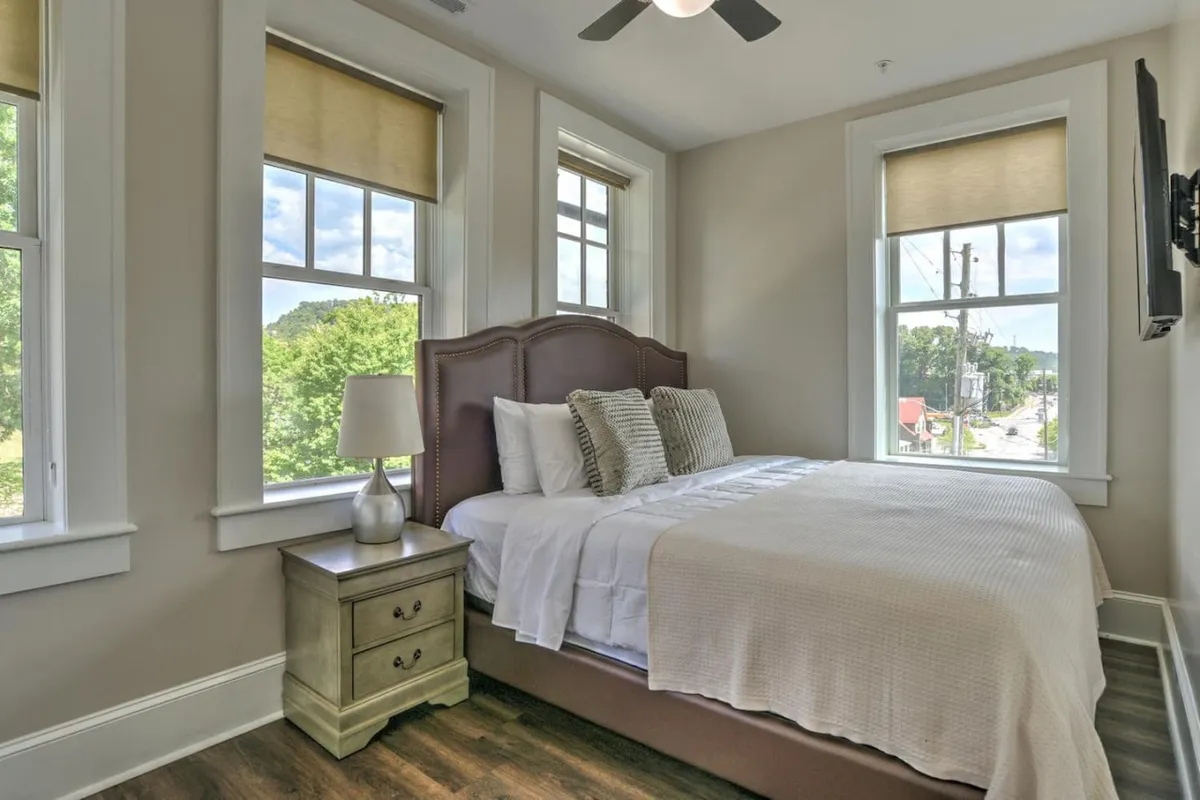 A serene bedroom featuring a plush bed centered against a wall with windows allowing natural light to flow in. A nightstand with a lamp and a television add comfort and convenience, while the overall palette and arrangement create a restful, inviting atmosphere.